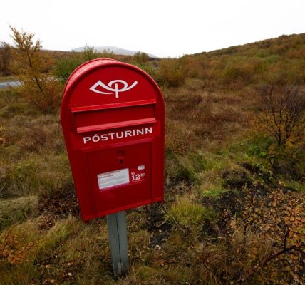 A red mailbox stands outdoors amidst autumn scenery.