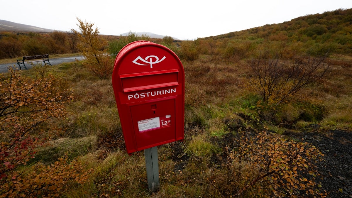 A red mailbox stands outdoors amidst autumn scenery.